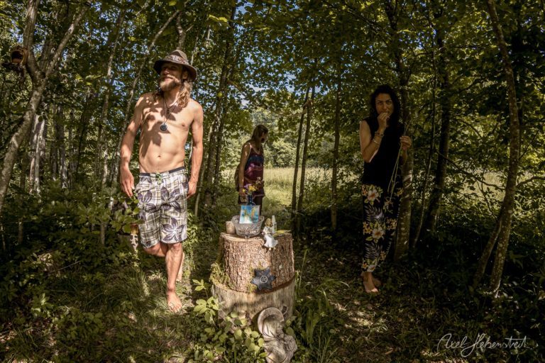 Vier Menschen in einem grünen Waldweg bei Sonnenschein, einer ohne Hemd mit Hut neben einem Baumstumpf-Altar – © Axel Hebenstreit