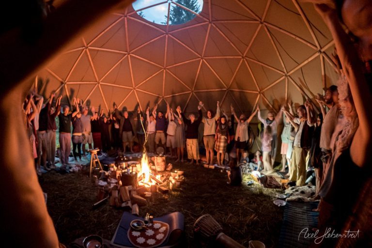 Lagerfeuer-Gemeinschaft unter geodätischer Kuppel mit Teilnehmern in festlicher Atmosphäre – © Axel Hebenstreit