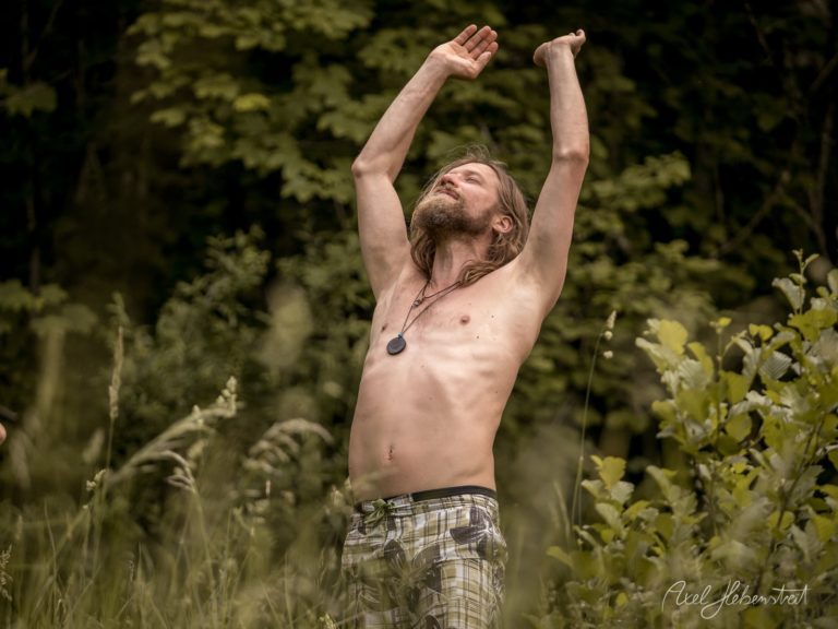 Mann mit langen Haaren und Bart reckt die Arme triumphierend zum Himmel in üppiger Waldlandschaft – © Axel Hebenstreit