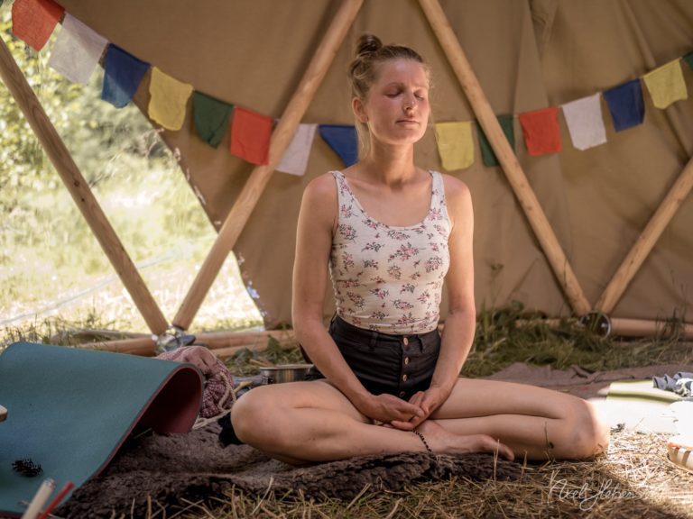 Frau in meditativer Pose sitzt auf Stroh in einem Glamping-Zelt mit bunten Gebetsfahnen – © Axel Hebenstreit