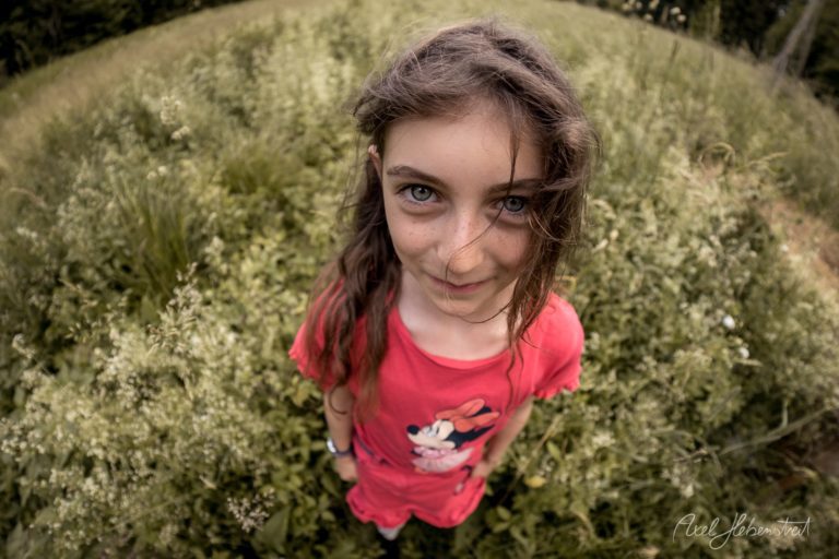 Junges Mädchen in rotem Shirt lächelt in die Kamera, umgeben von grünen Sträuchern in der Natur – © Axel Hebenstreit