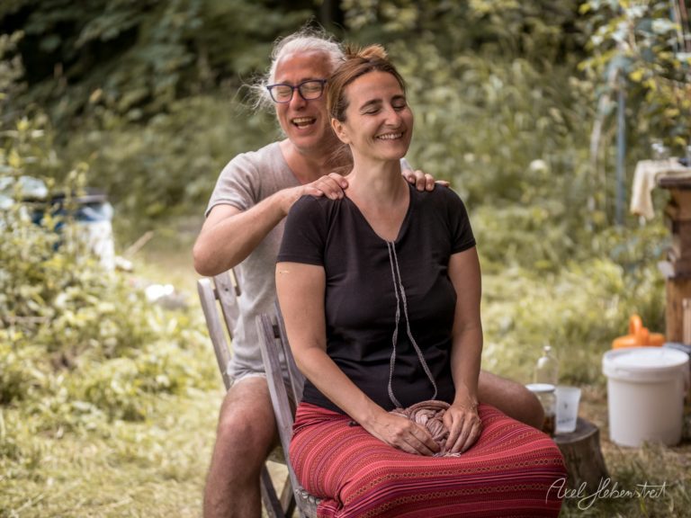 Glückliches Paar sitzt lachend in einem grünen Garten, umgeben von Pflanzen und Campingutensilien – © Axel Hebenstreit