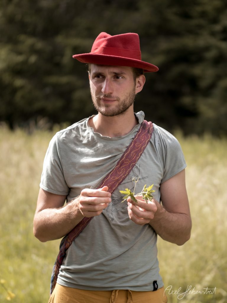 Mann mit rotem Hut hält Wildkräuter in einer grünen Wiese vor Waldhintergrund – © Axel Hebenstreit