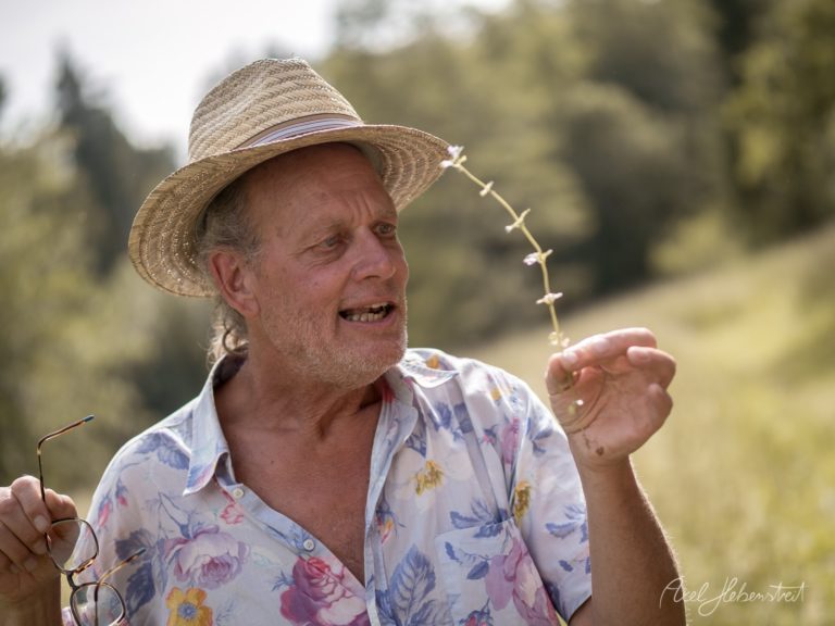 Älterer Mann mit Strohhut hält lächelnd eine blühende Pflanze in einer grünen Landschaft – © Axel Hebenstreit