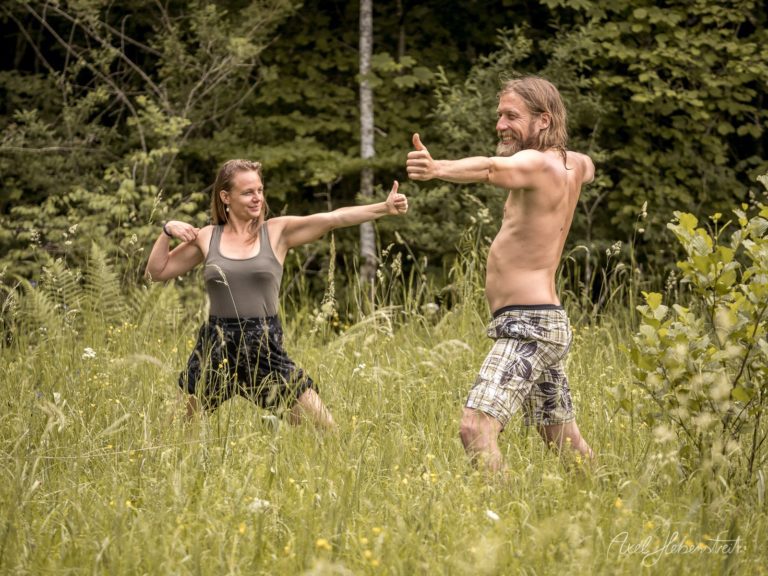 Zwei Menschen praktizieren Yoga in einer blütenreichen Wiese vor Waldkulisse mit ausgestreckten Armen – © Axel Hebenstreit