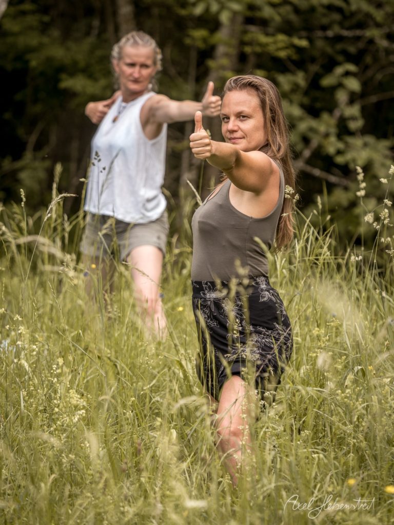 Zwei Frauen geben Daumen-hoch-Gesten in einem grünen Wildkräuter-Feld mit Wald im Hintergrund – © Axel Hebenstreit