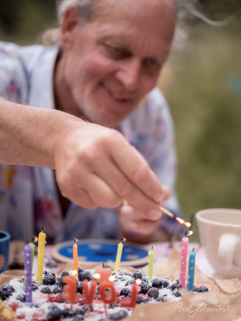 Ein Mann zündet eine Kerze auf einer bunten Geburtstagstorte mit Blaubeeren an – © Axel Hebenstreit