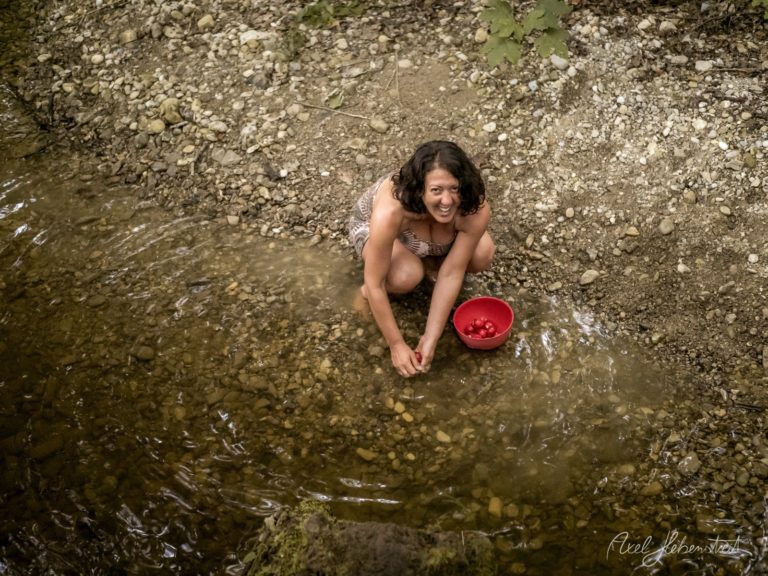 Frau sitzt fröhlich in einem flachen Gebirgsbach mit rotem Behälter und sammelt Wasser – © Axel Hebenstreit