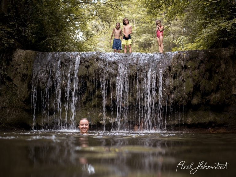 Eine Frau schwimmt vor einem kleinen Wasserfall, während drei Kinder amUfer stehen und lachen – © Axel Hebenstreit