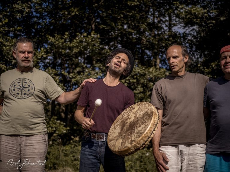 Eine Gruppe von vier Männern steht im Freien, während einer von ihnen Trommel spielt und singt. – © Axel Hebenstreit