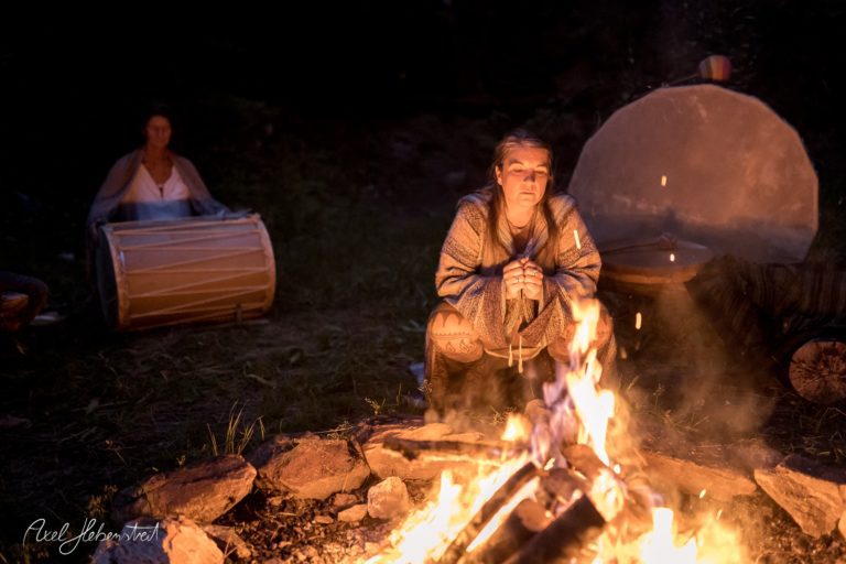Frau im Vordergrund meditiert am Lagerfeuer, während im Hintergrund Trommeln zu hören sind – © Axel Hebenstreit