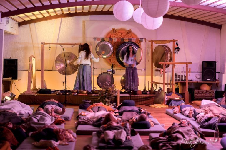 Relaxte Teilnehmer liegen auf Matten, während zwei Frauen mit großen Gongs in einem stimmungsvollem Raum musizieren. – © Axel Hebenstreit