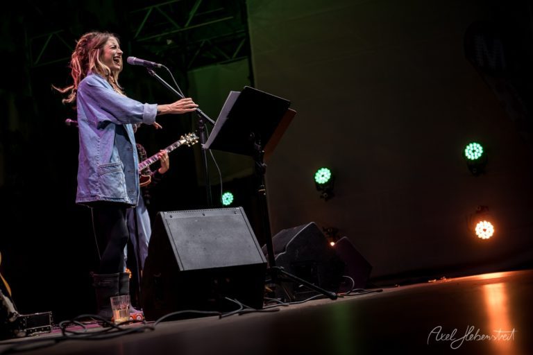 Eine Sängerin mit Mikrofon auf der Bühne lächelt, während sie zu einem Publikum auf einem Festival auftritt – © Axel Hebenstreit