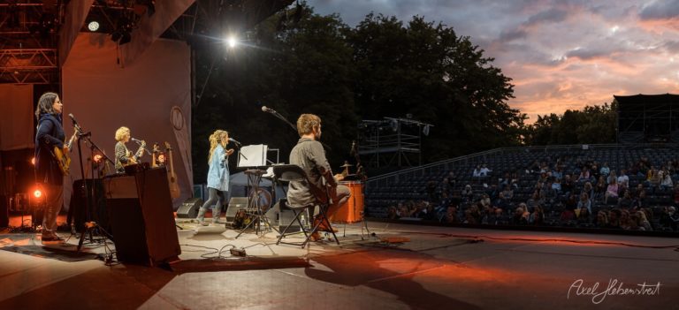 Musiker spielen auf einer Bühne bei Sonnenuntergang vor einem sitzenden Publikum in einer Freiluftarena – © Axel Hebenstreit