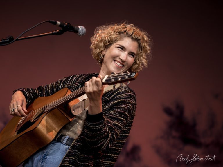 Frau mit Locken lächelt, während sie auf einer Bühne Gitarre spielt – © Axel Hebenstreit
