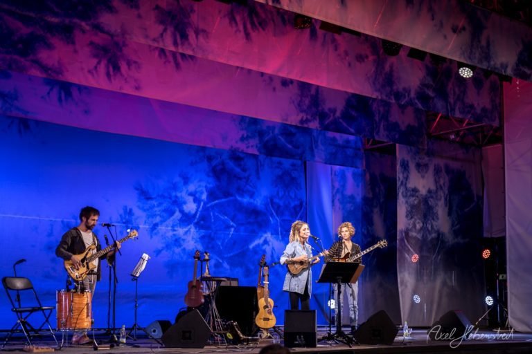 Zwei Sängerinnen performen auf einer Bühne mit buntem Hintergrund, begleitet von einem Musiker am Bass – © Axel Hebenstreit