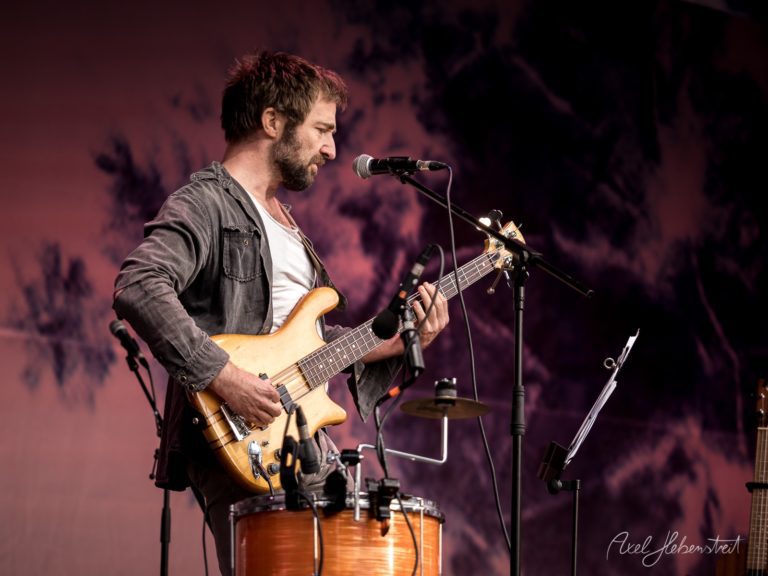 Musiker spielt Bass und singt auf einer Bühne bei einem Festival im Freien – © Axel Hebenstreit