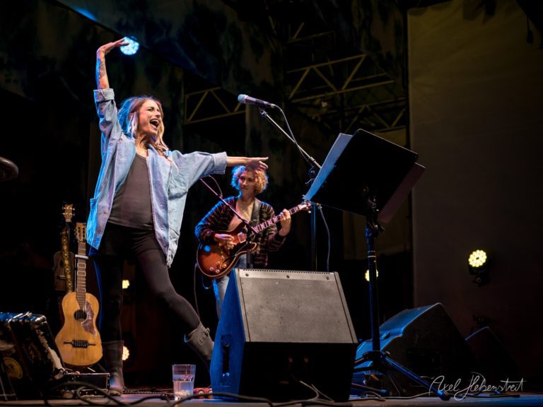 Eine Sängerin mit fröhlicher Ausstrahlung performt energisch auf der Bühne, während ein Musiker im Hintergrund spielt – © Axel Hebenstreit.