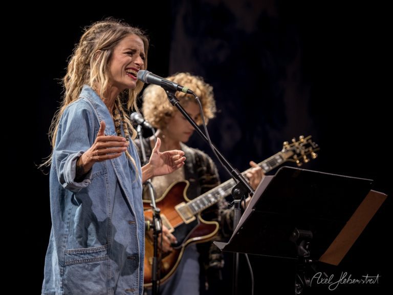 Frau singt leidenschaftlich in einem Konzert, während ein Gitarrist im Hintergrund spielt – © Axel Hebenstreit