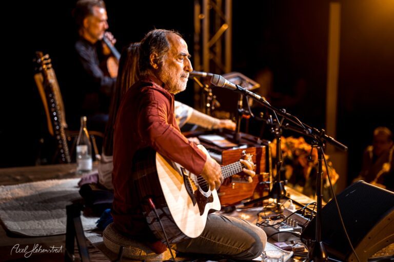 Ein Musiker mit Gitarre und Mikrofon spielt leidenschaftlich auf der Bühne bei einem Konzert – © Axel Hebenstreit