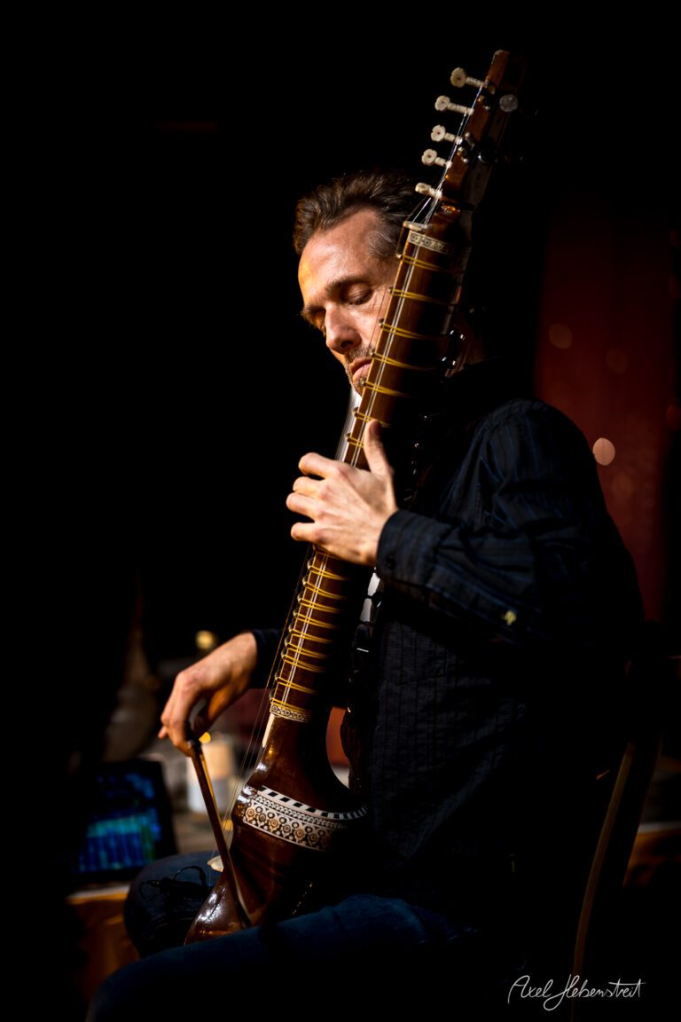 Ein Musiker spielt konzentriert auf einer Sitar, umgeben von warmem Bühnenlicht – © Axel Hebenstreit