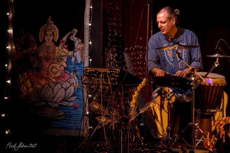 Ein Musiker spielt Perkussion vor einem kunstvoll gestalteten Hintergrund mit einer deity-Darstellung – © Axel Hebenstreit