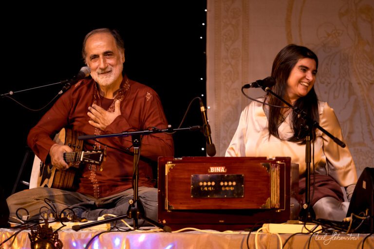 Zwei Performer sitzend auf der Bühne, lächeln und spielen Musik mit Gitarre und Harmonium – © Axel Hebenstreit