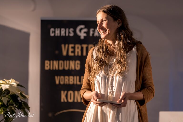 Eine lächelnde Frau steht auf der Bühne und hält ein Mikrofon, während sie eine Ansprache hält – © Axel Hebenstreit.