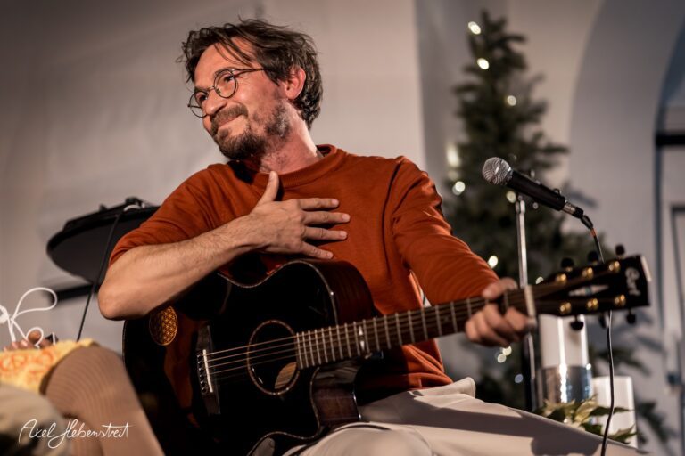 Musiker mit Gitarre lächelt nachdenklich, während er auf einer Bühne in festlicher Atmosphäre spielt – © Axel Hebenstreit