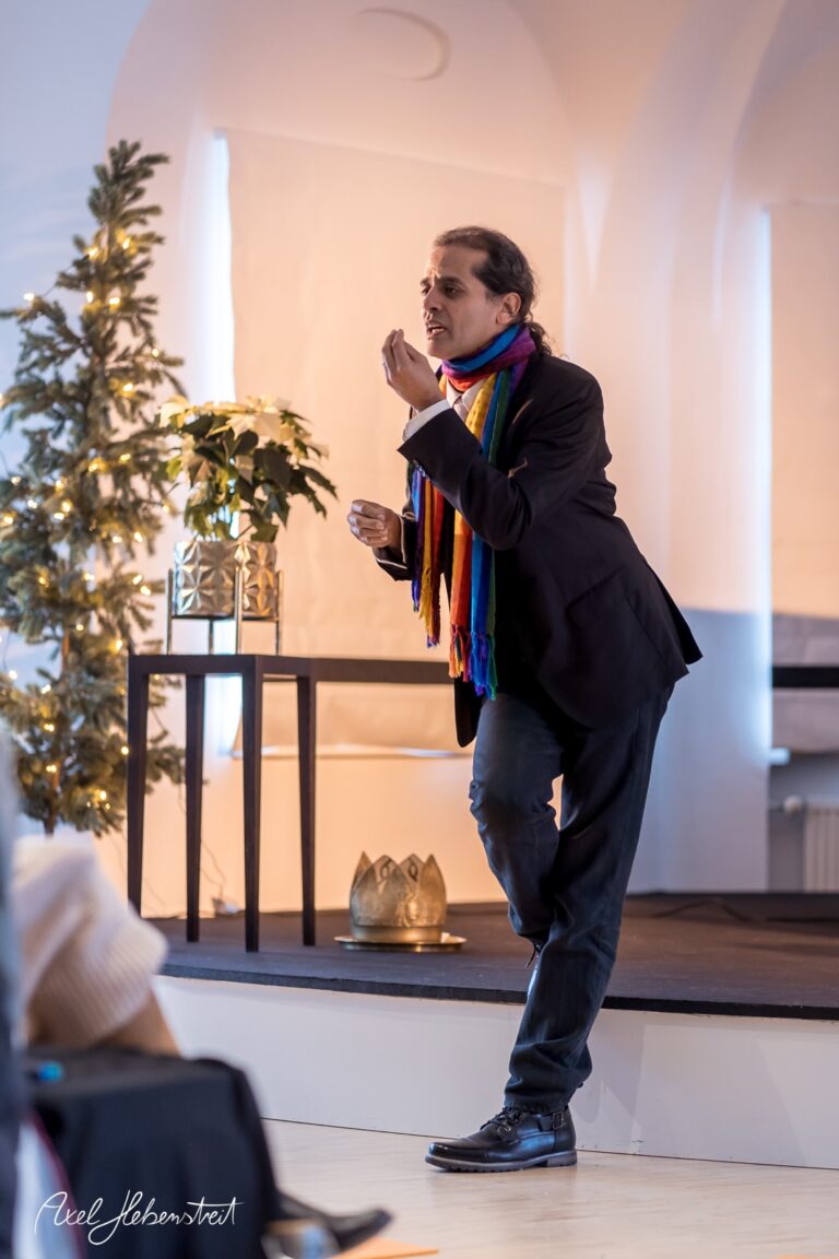 Ein Redner mit buntem Schal erklärt etwas leidenschaftlich in einem hellen Raum mit Weihnachtsdekoration – © Axel Hebenstreit