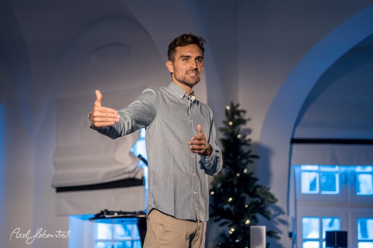Ein lächelnder Mann spricht engagiert auf einer Veranstaltung in einem festlich dekorierten Raum – © Axel Hebenstreit