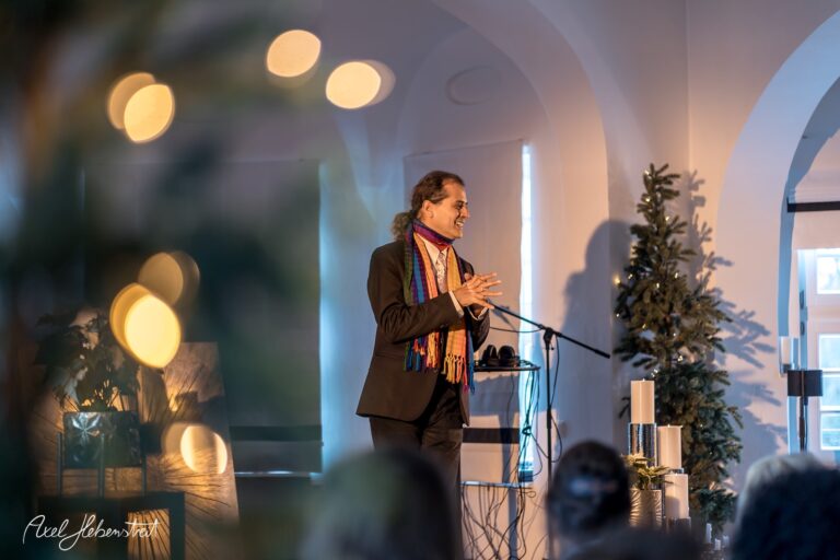 Ein Redner mit buntem Schal spricht mit Begeisterung auf einer Veranstaltung in einem festlich dekorierten Raum – © Axel Hebenstreit