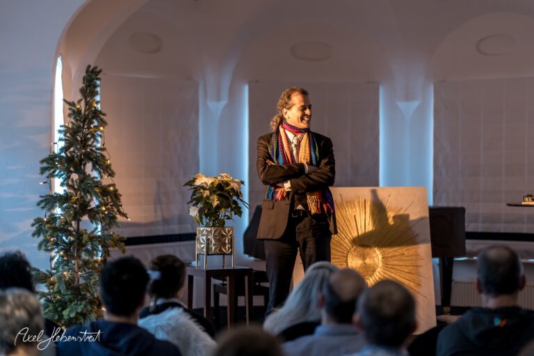 Ein Redner steht lächelnd auf der Bühne vor einem Publikum in einem festlichen, hell erleuchteten Raum – © Axel Hebenstreit