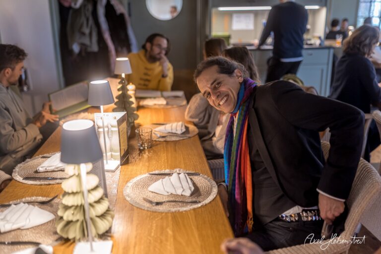Ein Mann mit buntem Schal lächelt an einem festlich gedeckten Tisch in einer einladenden Restaurantatmosphäre. – © Axel Hebenstreit