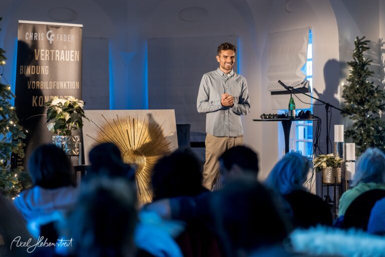 Ein lächelnder Referent steht auf der Bühne und hält einen Vortrag vor einem Publikum in einem modernen Veranstaltungsraum – © Axel Hebenstreit