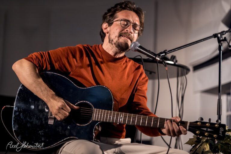 Ein Musiker in einem roten Pullover spielt leidenschaftlich Gitarre und singt auf einer kleinen Bühne. – © Axel Hebenstreit