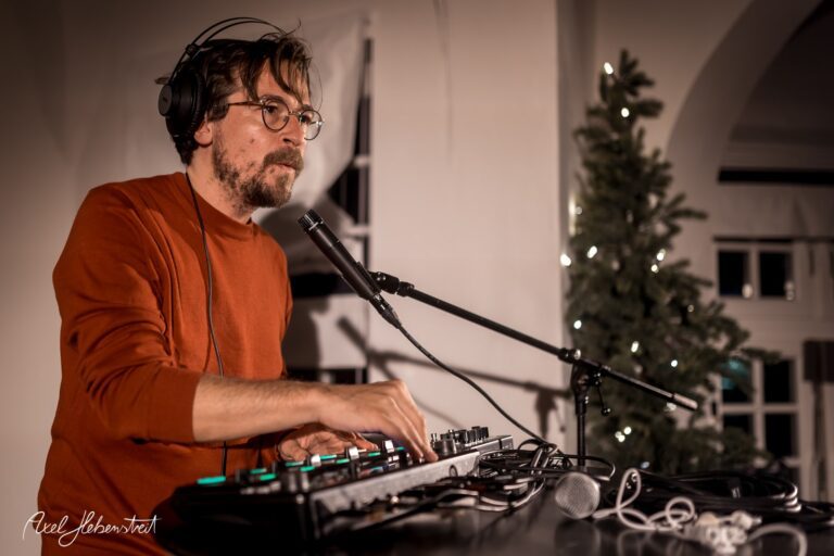 Ein DJ in einem gemütlichen Raum kreiert Musik, umgeben von sanfter Beleuchtung und einem Weihnachtsbaum. – © Axel Hebenstreit