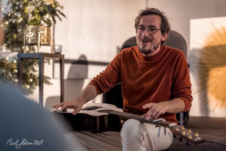 Ein leidenschaftlicher Musiker spielt auf seiner Gitarre und lächelt während einer intimen Atmosphären-Performance. – © Axel Hebenstreit