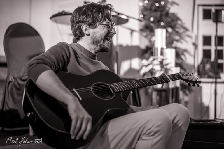 Lächelnder Musiker sitzt mit akustischer Gitarre in festlicher Atmosphäre – © Axel Hebenstreit