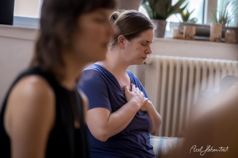 Eine Frau meditiert mit geschlossenen Augen und einer Hand auf der Brust in einem hellen Raum – © Axel Hebenstreit