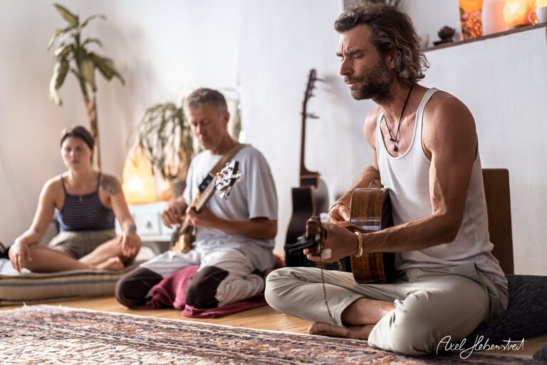 Musiker spielt Gitarre in einem entspannenden Raum, während zwei Zuhörer in meditativer Haltung lauschen – © Axel Hebenstreit
