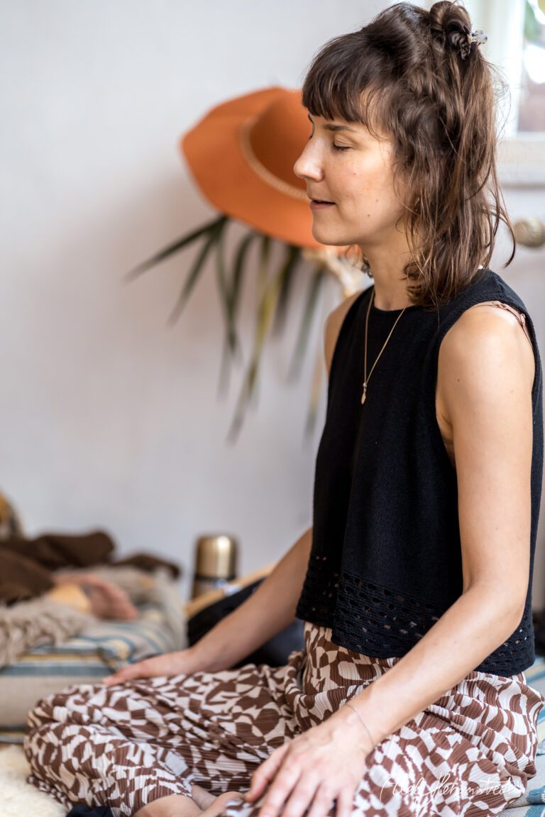 Junge Frau sitzt meditativ in einem stylischen Raum, umgeben von Pflanzen und gemütlicher Einrichtung – © Axel Hebenstreit