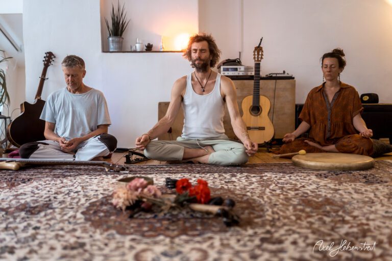 Drei meditierende Personen in einem harmonischen Raum, umgeben von Instrumenten und einem dekorativen Teppich – © Axel Hebenstreit