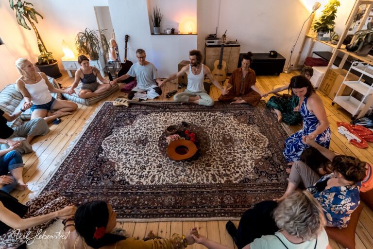 Eine Gruppe von Menschen sitzt im Kreis auf einem Teppich und praktiziert gemeinsam Yoga in einem hellen Raum – © Axel Hebenstreit