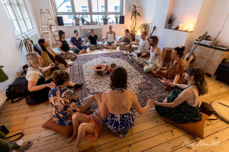 Eine Gruppe von Menschen sitzt im Kreis auf dem Boden, umgeben von Pflanzen, in einer entspannten Atmosphäre – © Axel Hebenstreit