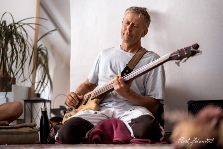 Ein Mann spielt entspannt auf einem E-Bass, umgeben von einer gemütlichen Atmosphäre. – © Axel Hebenstreit