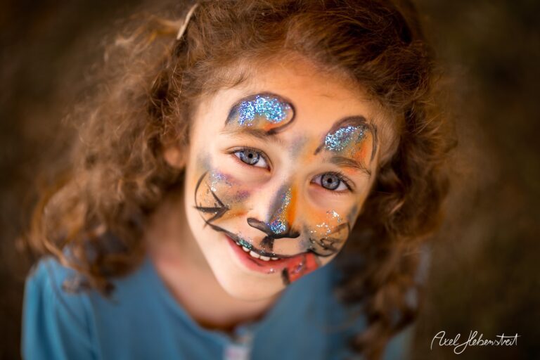 Ein Mädchen mit glitzerndem Katzen-Make-up lächelt fröhlich in die Kamera – © Axel Hebenstreit