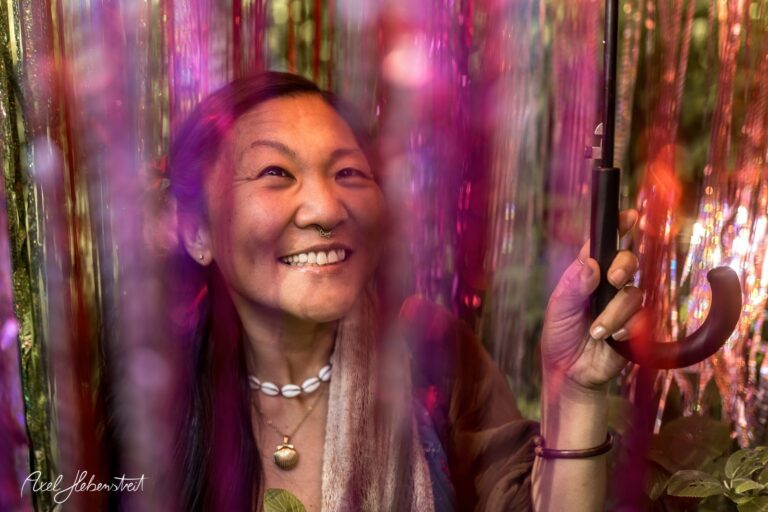 Frau mit lächelndem Gesicht und Regenschirm steht zwischen bunten, glitzernden Vorhängen – © Axel Hebenstreit