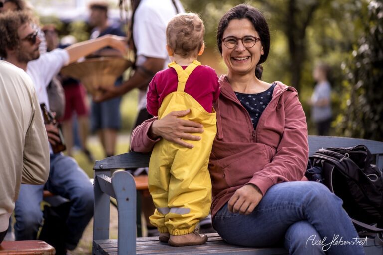 Eine lächelnde Frau sitzt auf einer Bank, während ein Kind in gelber Kleidung an ihr lehnt – © Axel Hebenstreit