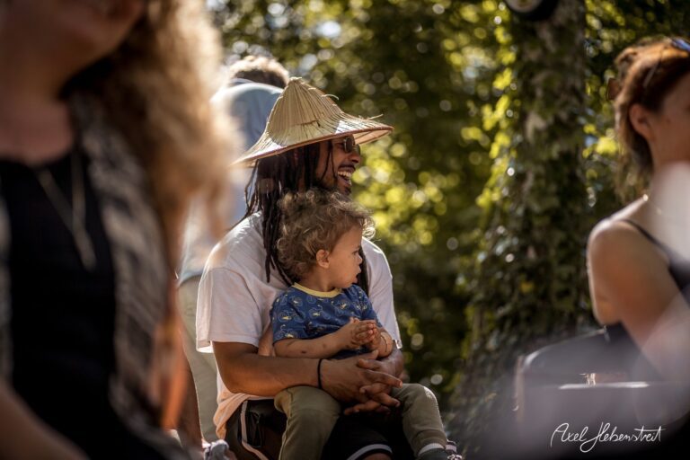 Ein fröhlicher Mann mit Strohhut sitzt im Freien und hält ein kleines Kind auf dem Schoß. – © Axel Hebenstreit